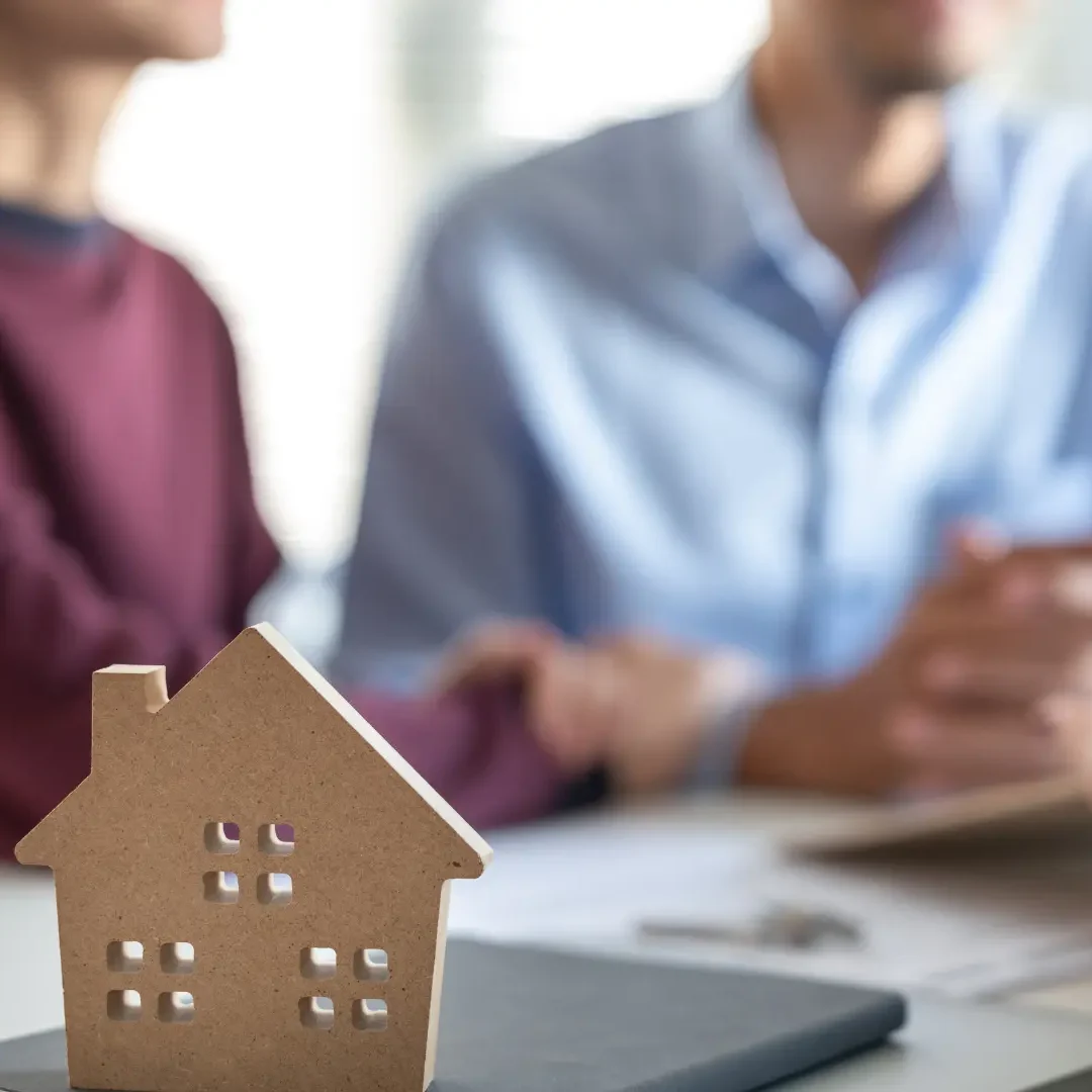 local-homebuyer-with-family Small wooden model house on a table with two siblings having a real estate discussion in the background about an inherited house, focusing on property consultation. The woman wears a burgundy sweater and the man wears a light blue shirt, both seated at a white table with documents